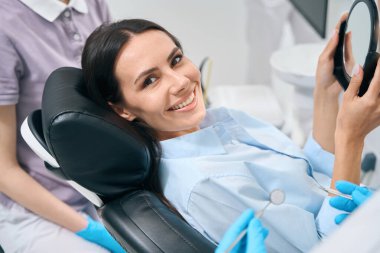 Çekici dişçi kadın hasta kameraya sağlıklı dişli bir gülümsemeyle bakıyor. Doktorun elinde ayna tutmasından memnun. Özel stomatoloji kliniği.