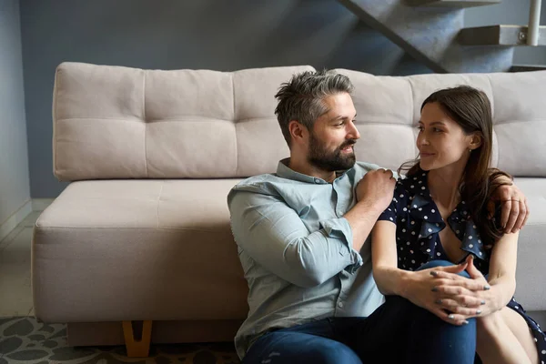 Man and a woman are sitting on the floor by the sofa, husband put his arm around his wifes shoulders