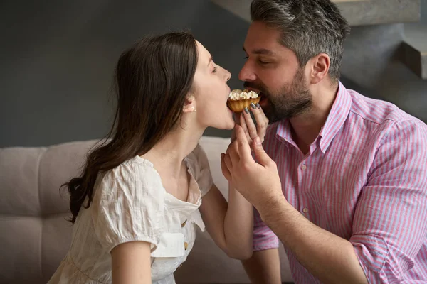 Man and a woman sit on the couch and at the same time bite off a cake, they feel good together