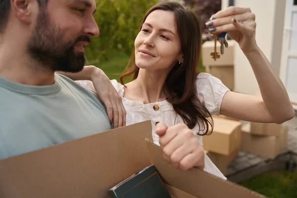 Attractive woman showing keys and looking at her man who holding box in yard