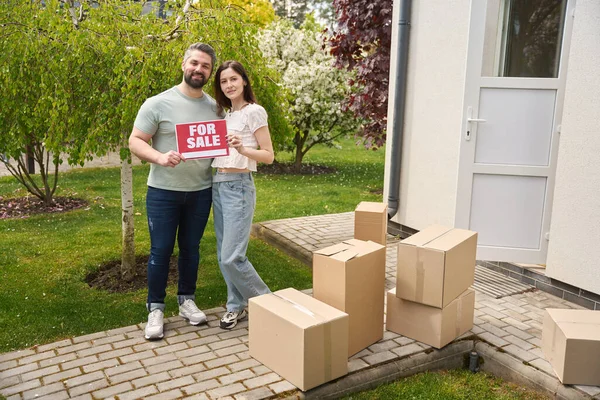 Young man and woman looking at camera and holding sign for sale while standing in yard