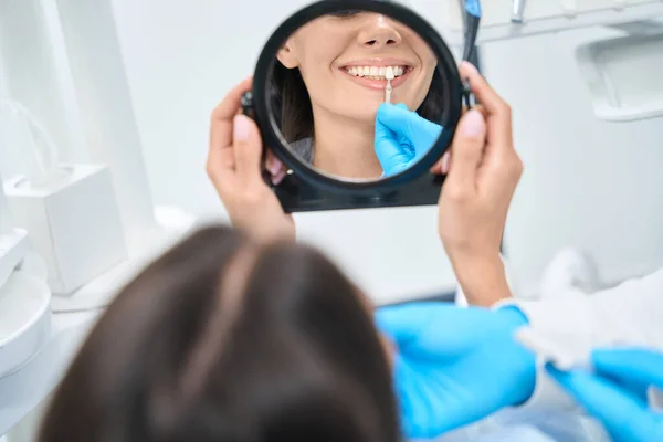 Female client coming at dentist appointment to fit veneers and choose needed color, concealing cosmetic imperfections of teeth, reflection in mirror