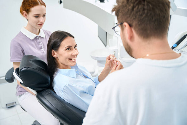 Smiling woman enjoying her new removable dental prosthesis sitting in orthodontic chair and listening to stomatologist and his nurse advices about wearing