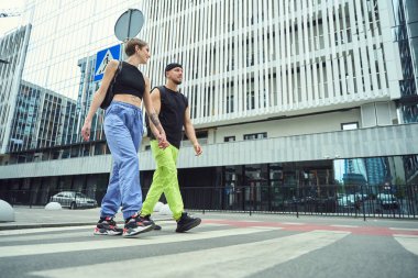 Kız arkadaşı olan bir adam, el ele tutuşuyor, yaya geçidi boyunca yürüyor, yol tabelasının yanında.