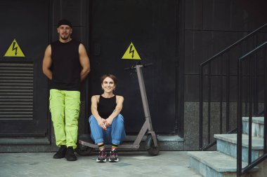 Birkaç gayri resmi vatandaş modern bir şehir binasının duvarında dinleniyor. Genç bir kadın bir scooter 'a binmiş.