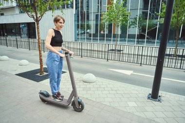 Spor giyimli bir kadın şehrin sokaklarında scooter sürüyor, zayıf ve kısa saçlı.