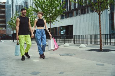 Adam ve genç bir kadın, modern bir binanın arka planında el ele yürüyorlar. Alışveriş torbaları var.