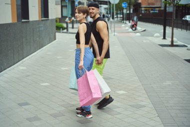 Genç adam ve kadın el ele tutuşarak yürüyor, kaldırımda, dişinin alışveriş torbaları var.