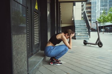 Üzgün genç bir kadın bir şehir binasının duvarının yanında oturuyor, yakınlarda bir scooter duruyor.