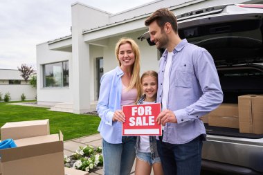 Genç kızı olan evli bir çift yeni evin avlusunda duruyorlar, eşleri satılık bir tabela tutuyorlar.