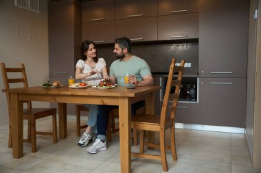 Karı koca, güzel bir mutfakta, ahşap bir masada kahvaltı ediyor. İnsanlar keyifli.