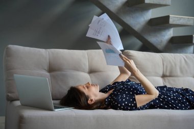 Puantiyeli bir elbise giymiş esmer kanepeye uzanıp belgelerle uğraşıyor. Yakınlarda bir laptop var.