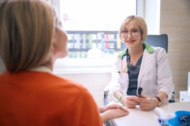 Turuncu kazaklı bir kadın bir çocuk doktoruyla iletişim kuruyor, kızı yanında oturuyor.