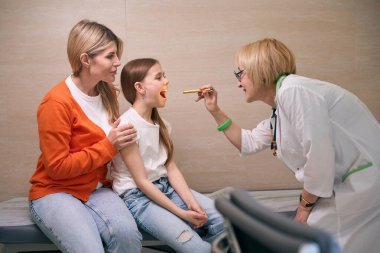 Klinikteki kadın çocuk doktoru, çocuğun annesinin yanındaki küçük bir hastanın boğazını muayene ediyor.