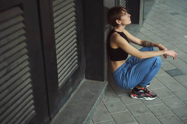 穿着运动服的年轻女子坐在一座城市建筑的墙边 剪短了头发 — 图库照片