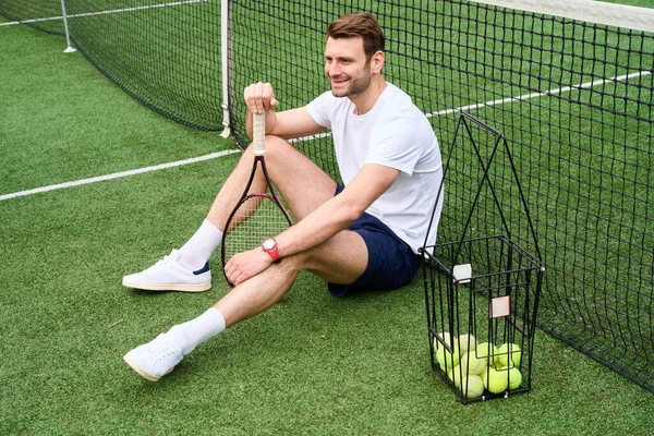 テニスコートの草の上に座っている笑顔のハンサムな男 彼はラケットとボールのバスケットを持っています — ストック写真