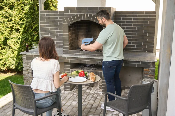 夫は裏庭の大きなオーブンで火を起こし 妻は近くの野菜をカットする — ストック写真