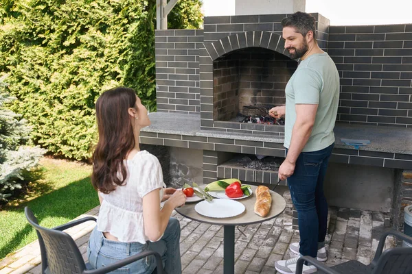 笑顔の男性は大きなオーブンで火を作ります 彼の隣に彼の妻は野菜をカット — ストック写真