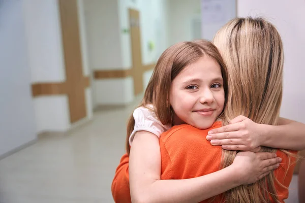 笑顔の少女は優しく彼女の母親を抱き クリニックでオレンジ色のセーターでブロンド — ストック写真