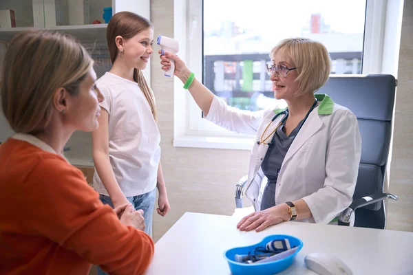 小児オフィスの女性小児科医は オレンジ色のセーターの少女の母親の体温を測定する — ストック写真