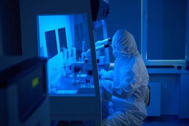 Steril üniformalı bir laboratuvar çalışanı mikro manipülatör seti kullanır, ayrıca güçlü bir mikroskop kullanır.