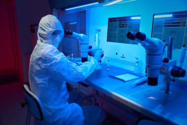 Koruyucu üniformalı embriyolog ICSI prosedürü uyguluyor, mikro manipülatör seti ve güçlü mikroskop kullanıyor.