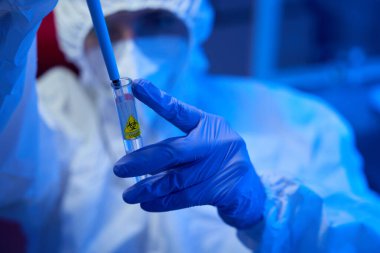 Steril odada elinde tehlike sembolü olan bir test tüpü tutan bir adam var. Laboratuvar asistanı koruyucu eldivenlerle çalışıyor.