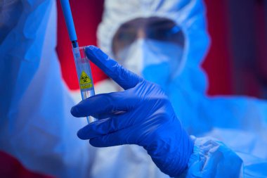 Steril üniformalı bir adam elinde tehlike sembolü olan bir test tüpü tutuyor. Laboratuvar asistanı koruyucu eldivenlerle çalışıyor.
