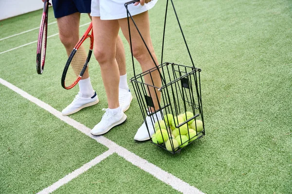 テニスウェアのパートナーはテニスコートに沿って歩き 女性はテニスボールを持つバスケットを持っています — ストック写真