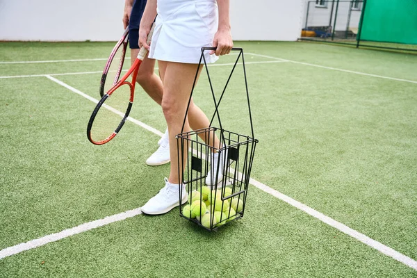 テニスパートナーはラケットとテニスコートに沿ってボールのバスケットを歩き 裁判所は高い塀で囲まれています — ストック写真