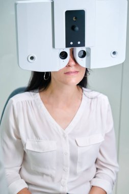 Waist-up portrait of female patient looking through electronic phoropter lenses while sitting on examination chair clipart