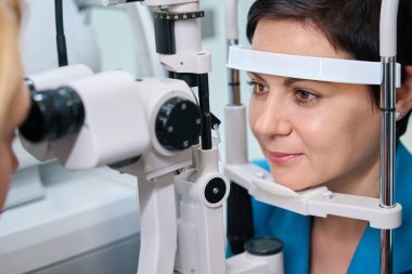 Optometrist tarafından yapılan göz muayenesi sırasında çenesi ve alnı desteklenirken kadın hasta kesik lambayla karşı karşıya.