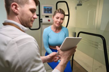 Çalışkan fitness eğitmeni, modern odada uzun saçlı bayan müşterisine tablet gösteriyor.