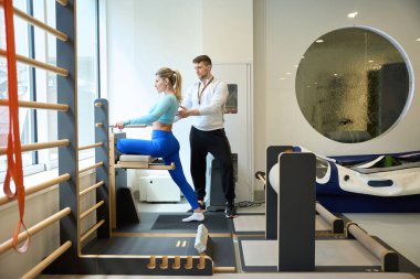 Uzun saçlı genç bir bayan spor yaparken beden eğitimi hocası onu modern spor salonunda izliyor.