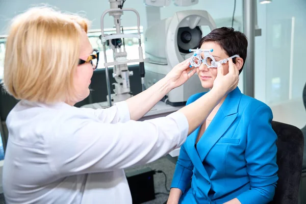 女性の眼科医は視力スクリーニングの前に顧客の表面に試験レンズ フレームを置きます — ストック写真