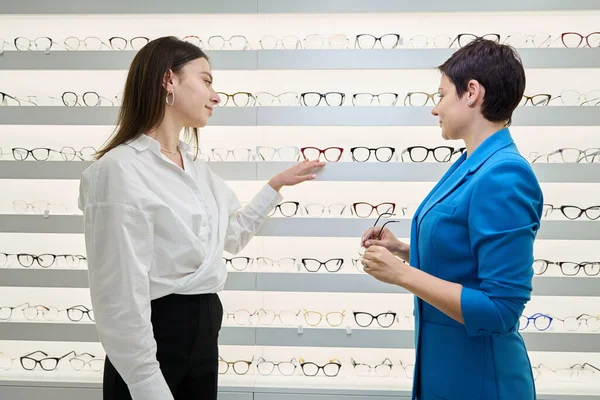 Female optician demonstrating eyeglass frames on eyewear display shelf to adult woman