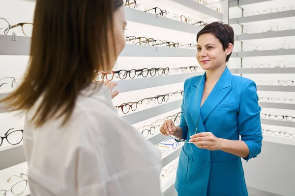 Optical dispenser showing spectacle frames on eyewear display shelf to smiling client