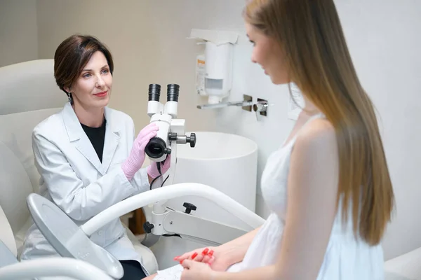 女性婦人科医は彼女の仕事で婦人科顕微鏡を使用し 若い女性は検査のために婦人科の椅子に位置しています — ストック写真