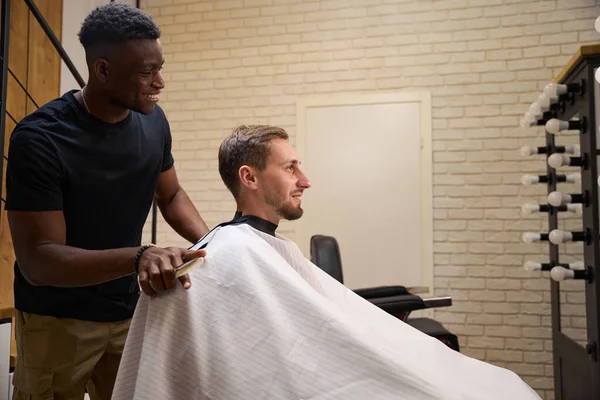 Young man sits in a barbers chair, an African American master chats with him in a friendly way