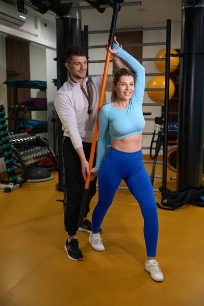 Smiling woman in fitness wear doing sport exercises while coach helping her in fitness center