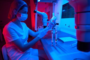 Koruyucu maskeli bir kadın laboratuvar asistanı bir ICSI prosedürü uyguluyor. Kriyo laboratuarında modern ekipmanlar.