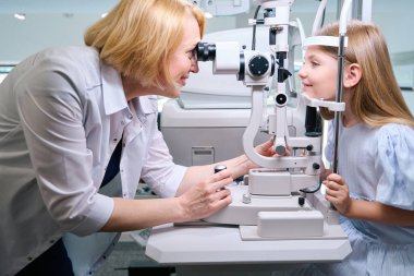 Neşeli kadın optometrist pediyatrik hasta gözlerine kesik lamba mikroskobundan bakıyor.