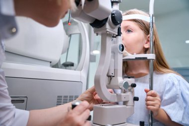 Deneyimli kadın optometrist, genç hasta gözlerini yüksek güçlü yarık lamba mikroskobuyla inceliyor.