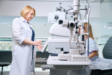 Focused female optometrist assessing refractive error in pediatric patient with automated refractor clipart
