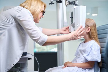 Gülümseyen dost canlısı bayan göz doktoru pediyatrik hastanın yüzüne dijital phoropter yerleştiriyor.