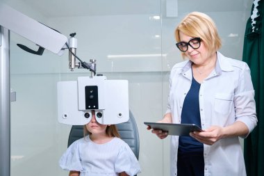 Little girl seated on examination chair looking through digital phoropter lenses in presence of ophthalmologist clipart