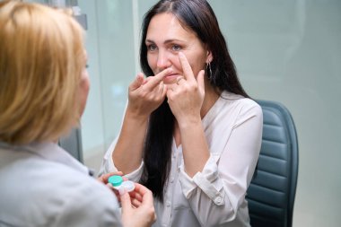 Yetişkin bir kadın, göz doktorunun önünde lens tutarken göz kapağını indiriyor.