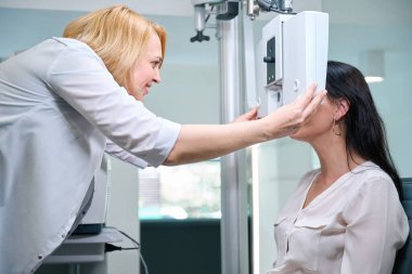 Smiling orthoptist placing electronic phoropter head in front of adult woman eyes clipart