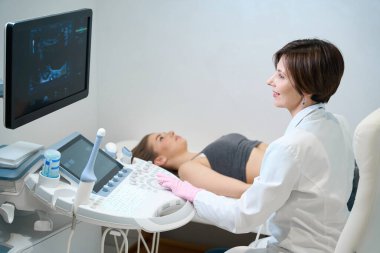 Dişi uzist hamile bir kadının cenini için ultrason prosedürü uygular, doktor gülümseyerek monitöre bakar.