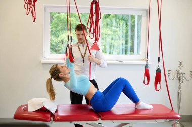 Güzel bayan müşteri içeride kırmızı kablo kullanarak antrenörle süspansiyon egzersizleri yapıyor.